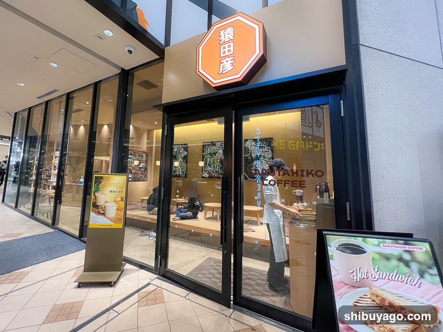 A modern coffee shop entrance with a prominent orange hexagonal sign displaying Japanese characters '猿田彦' (Saitaiko) above the door. Large glass windows reveal interior seating and staff in white uniforms preparing drinks. Two promotional stands outside advertise 'Hot Sandwich' and other menu items.