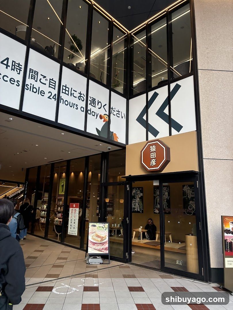 A modern shop entrance with large glass windows and doors, featuring signage reading '24 hours a day' in Japanese and English. A sign above the door displays '猿田彦'. Inside, customers are seated at tables.