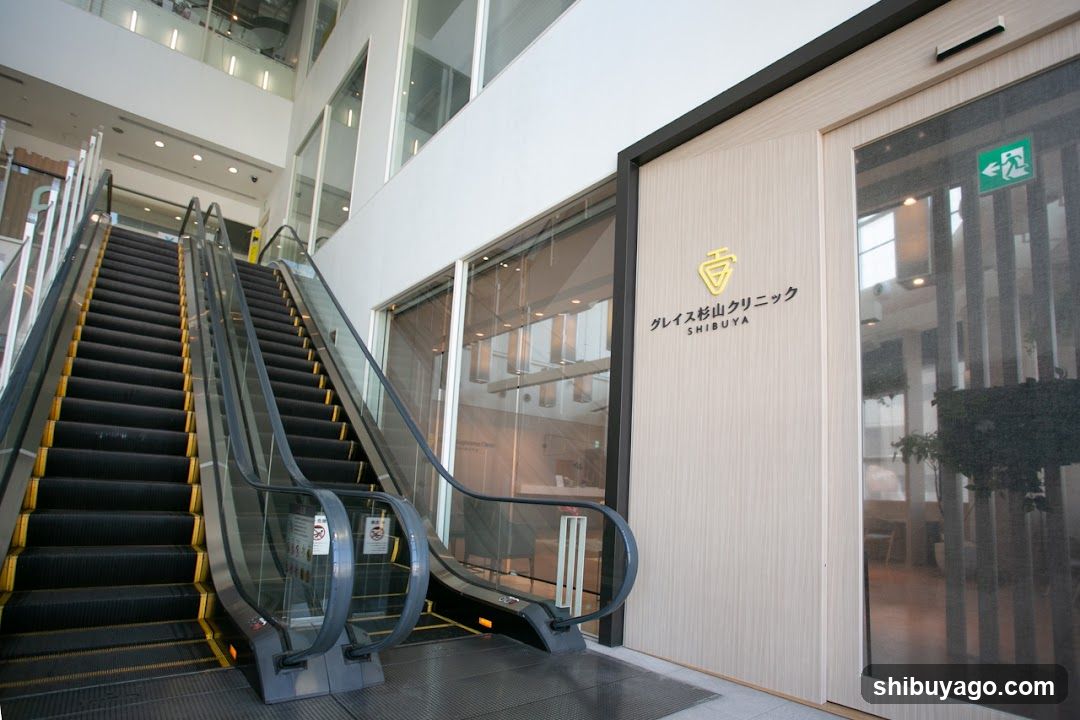 A modern building entrance featuring a set of escalators leading up to the ground floor. The facade includes large glass windows and doors, with signage reading 'グレイス杉山クリニック' (Grace Sugiya Clinic) in Japanese characters alongside English text.