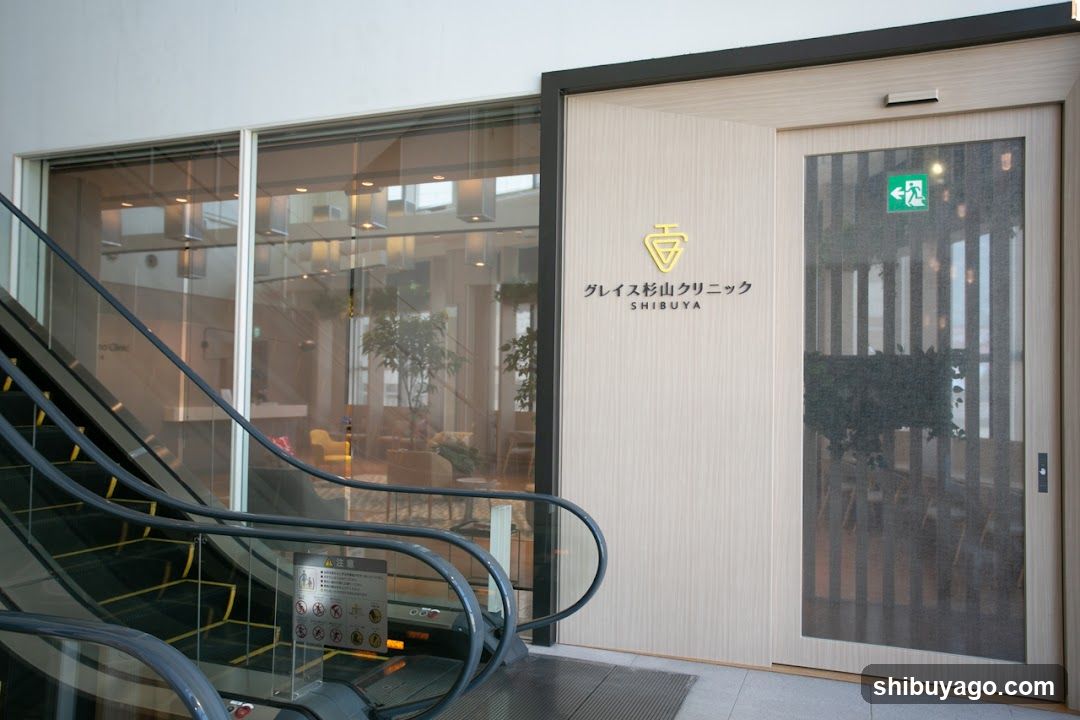 Exterior view of the Greys Shibuya Clinic entrance, featuring a modern glass facade with an escalator on the left and a wooden door with signage displaying 'グレイズ杉山クリニック SHIBUYA' in Japanese and English.
