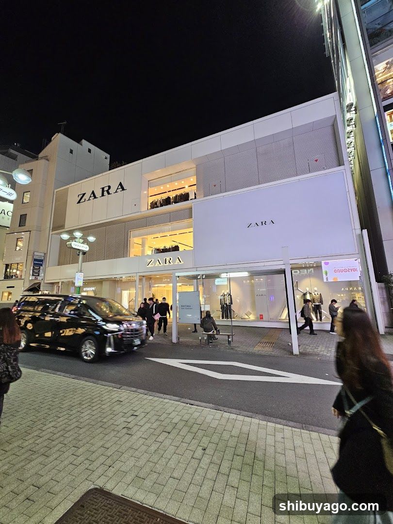 Night view of a Zara storefront with illuminated signage and large glass windows displaying mannequins. A black van is parked nearby, and pedestrians are walking on the sidewalk.