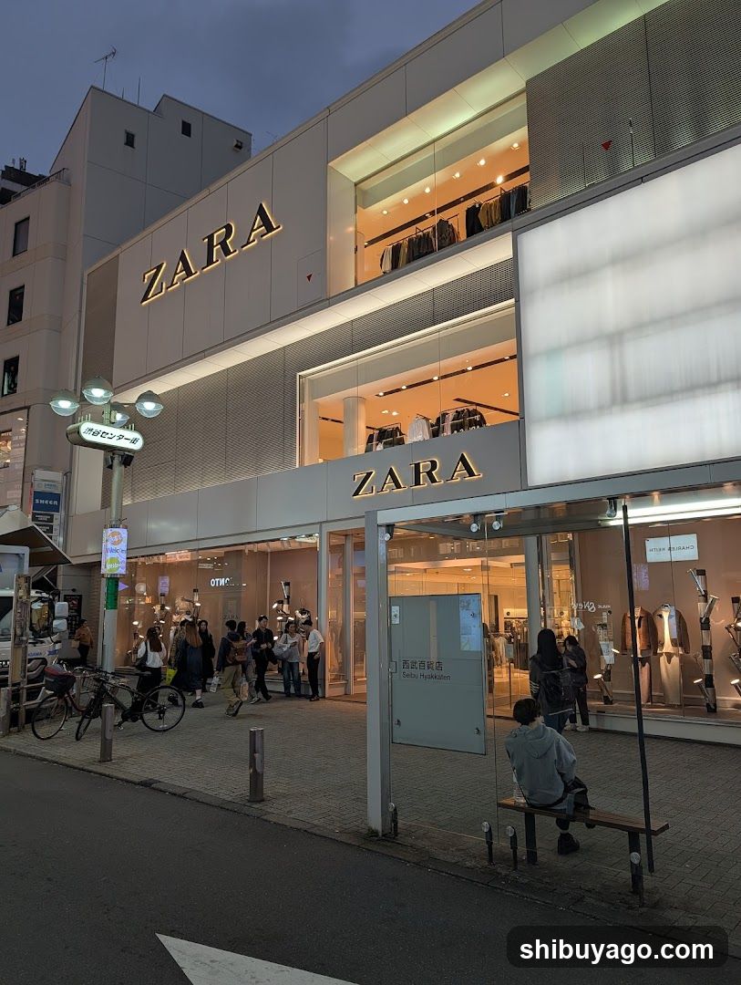 Evening shot of a Zara store facade with illuminated signage on upper and lower levels. Large glass windows display clothing racks inside. Pedestrians walk along sidewalk; one person sits on bench near entrance. Street lamp and bicycles visible nearby.