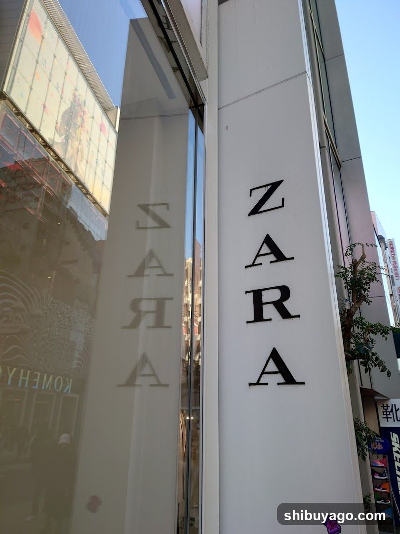 Photo of the Zara store facade showing vertical signage with 'ZARA' in bold black letters on a white pillar and etched lettering on glass windows. Reflections of surrounding buildings are visible.