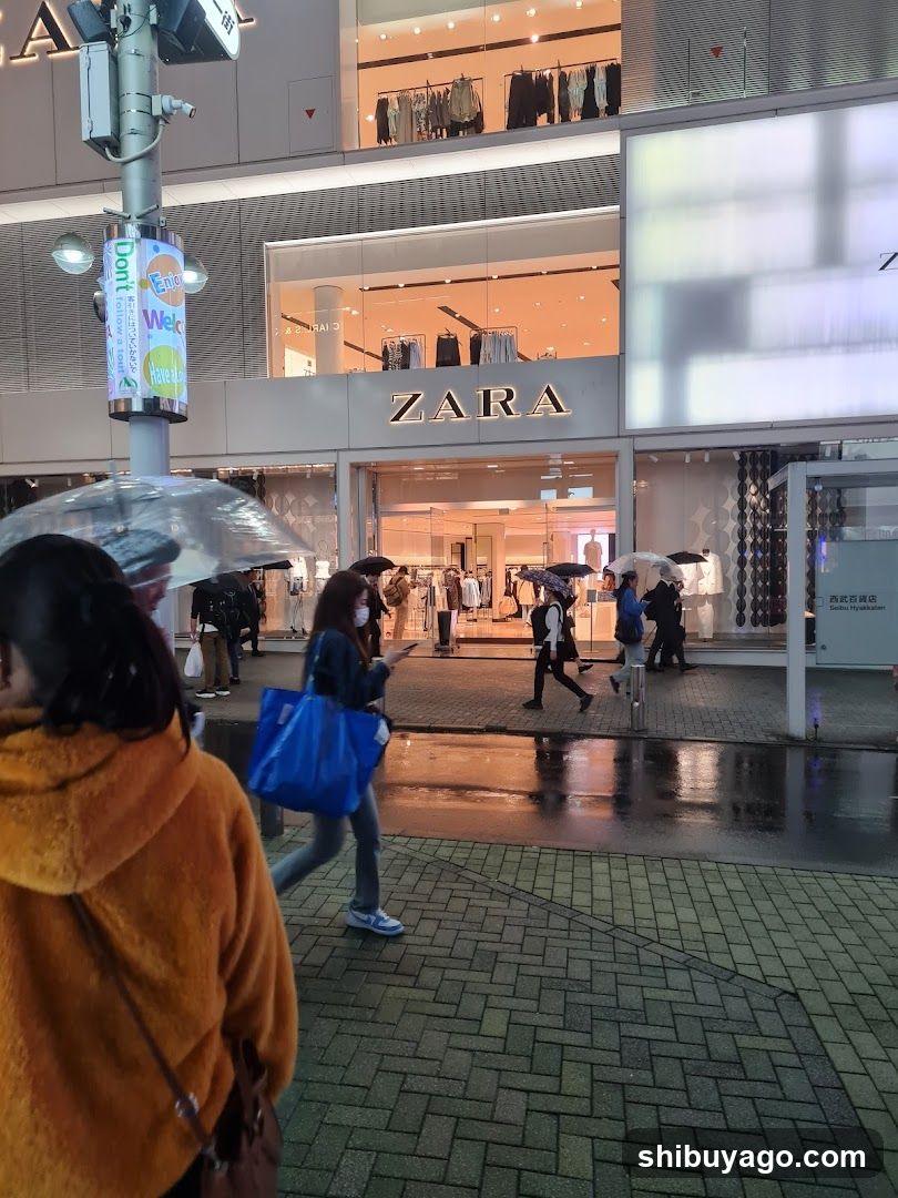 A photo of the physical exterior entrance and storefront facade of a Zara store. The building features large glass windows displaying clothing racks inside, with the 'ZARA' sign clearly visible above the entrance. People are walking on the sidewalk in front, some holding umbrellas, suggesting rainy weather.