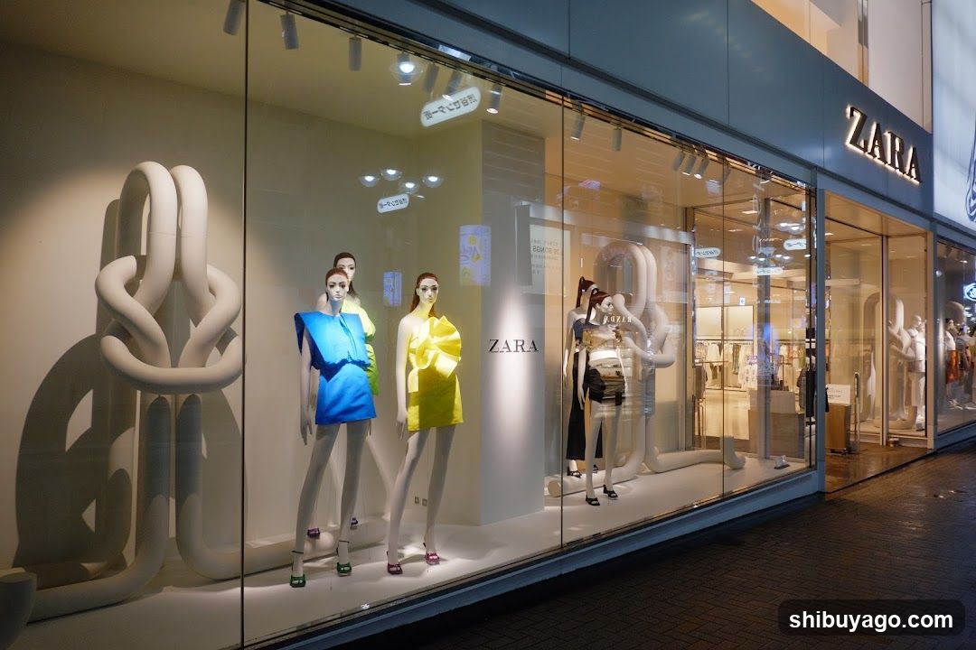 A Zara store window display featuring mannequins dressed in colorful outfits, including blue and yellow garments. A large abstract sculpture is visible on the left side of the window. The Zara logo is prominently displayed above the entrance.