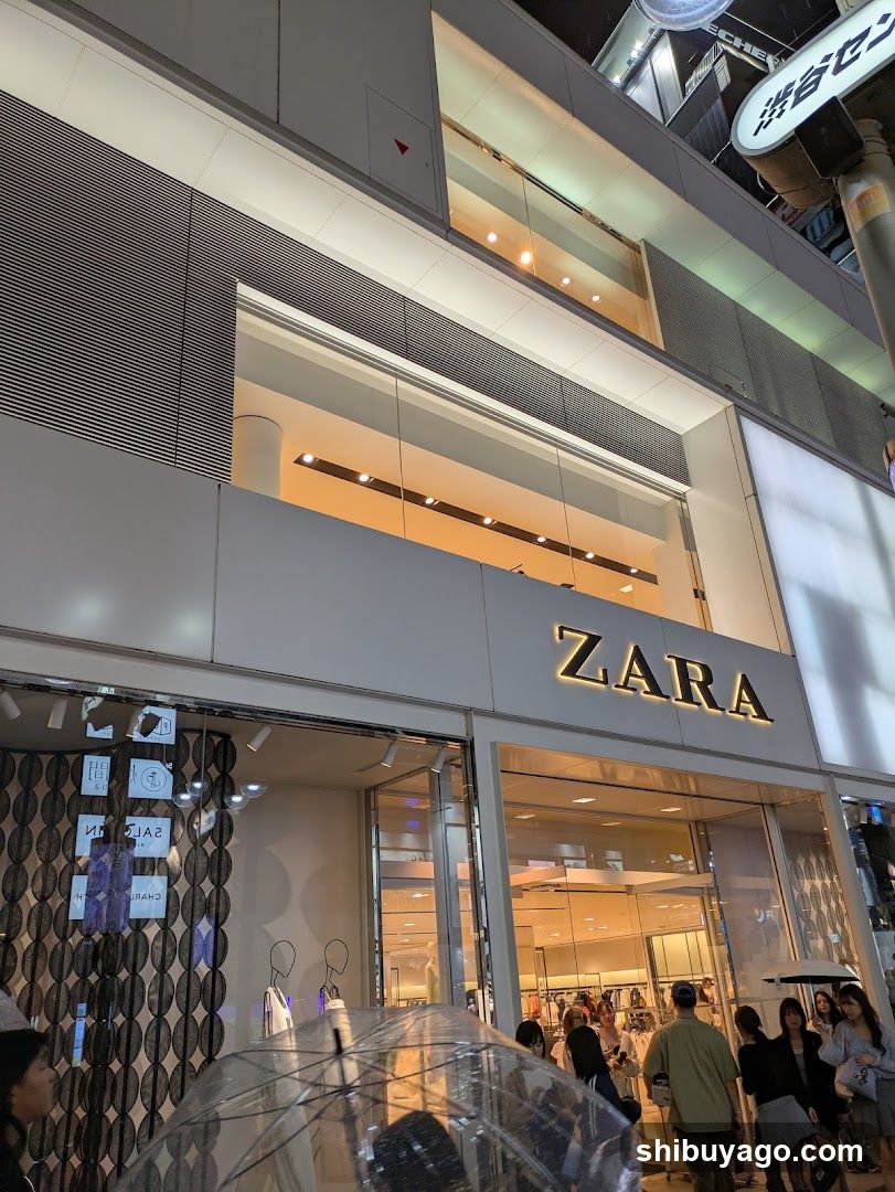 A modern Zara store facade with large glass windows and a prominent 'ZARA' sign above the entrance. The interior is visible through the glass, showing clothing racks and mannequins. People are gathered outside, some holding umbrellas, suggesting light rain.