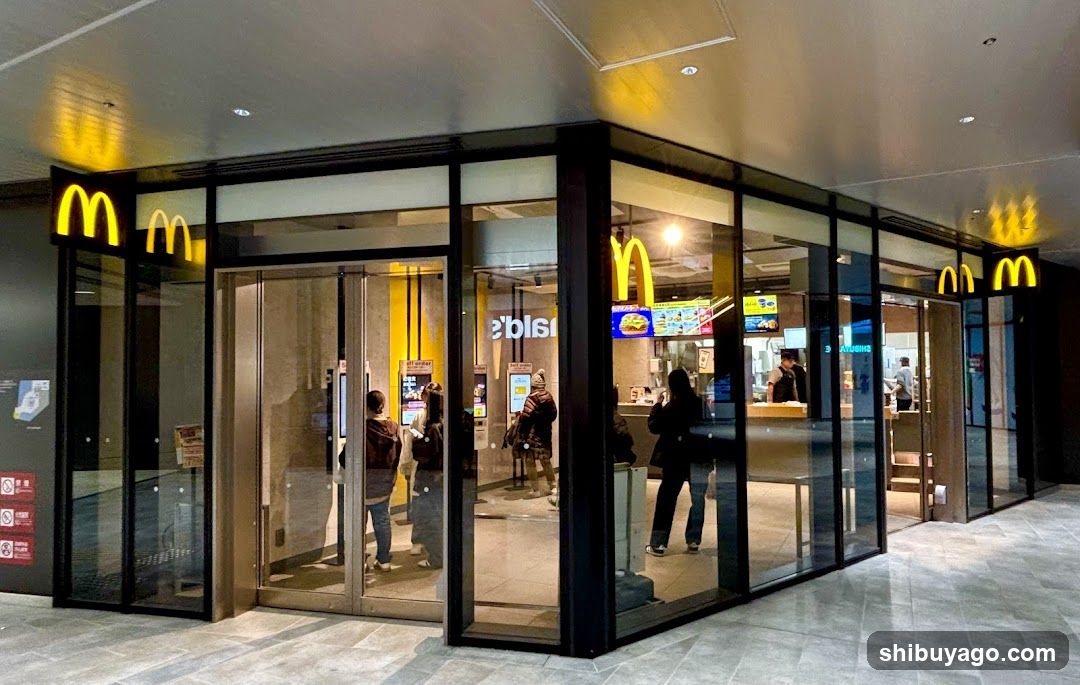 A modern McDonald's storefront with large glass windows and doors, featuring the iconic golden arches logo illuminated on both sides of the entrance. Inside, customers are visible near the counter and seating area.