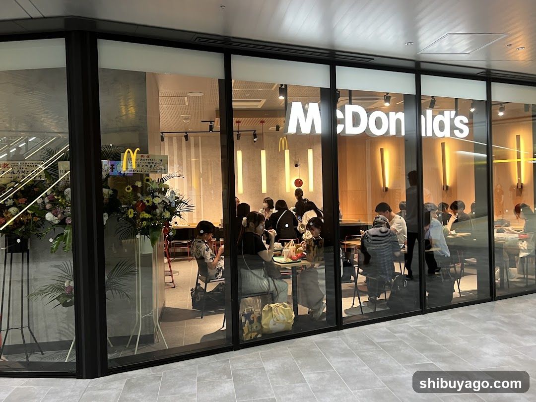 The image shows the exterior of a McDonald's restaurant, featuring large glass windows that offer a clear view into the dining area. Inside, customers are seated at tables enjoying their meals. The iconic golden arches logo is visible on the wall inside, and there are floral arrangements placed near the entrance. The lighting is warm, creating an inviting atmosphere.
