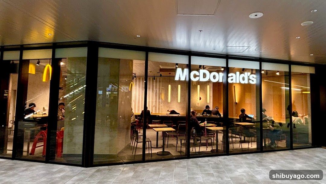 A modern McDonald's storefront with large glass windows and black frames. The illuminated 'McDonald's' sign is prominently displayed above the entrance. Inside, customers are seated at tables under warm lighting.