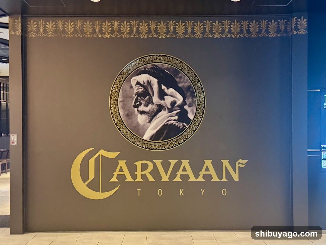 A wall-mounted sign featuring a circular portrait of an elderly man with a beard and head covering, surrounded by ornate golden detailing. Below the portrait is the word 'CARVAAN' in stylized gold lettering, followed by 'TOKYO' beneath it. The background is dark brown with decorative patterns along the top edge.