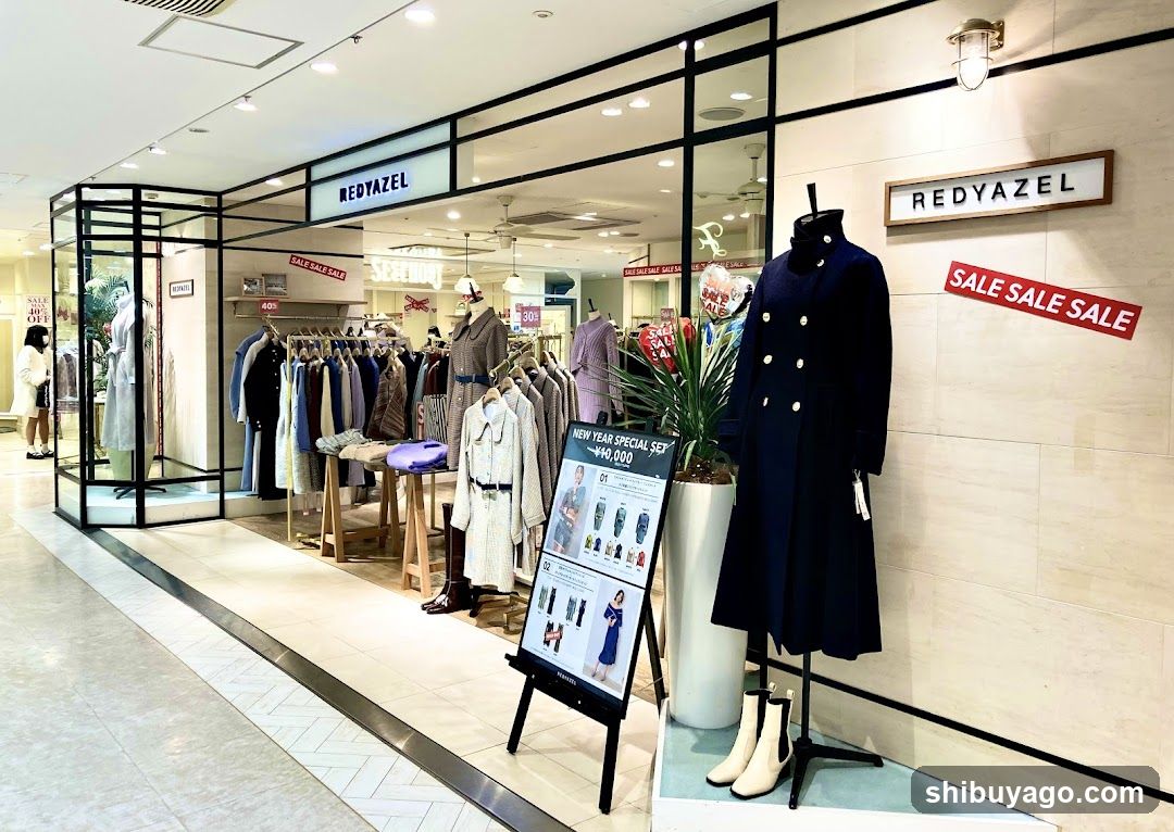 A modern retail store with glass facade and black metal framing. The sign 'REDYAZEL' is visible above the entrance. A large navy coat on a mannequin stands outside, next to a promotional stand displaying clothing items. Red banners reading 'SALE SALE SALE' are mounted on the wall.