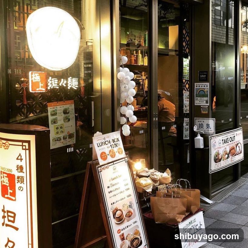 The image shows the exterior of a restaurant or shop, featuring illuminated signage on the building facade. A large circular light fixture is mounted above the entrance, accompanied by vertical Japanese characters. Several menu boards are placed outside, including one labeled 'LUNCH BOX' with prices and descriptions. Inside, shelves stocked with bottles are visible through glass doors. Paper bags and food items are displayed near the entrance.