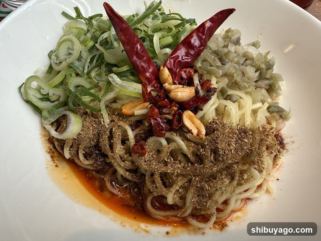A close-up of a bowl containing noodles topped with chopped green onions, dried red chilies, peanuts, and a generous amount of brown spice powder. The dish is served in a white bowl with visible oil at the bottom.