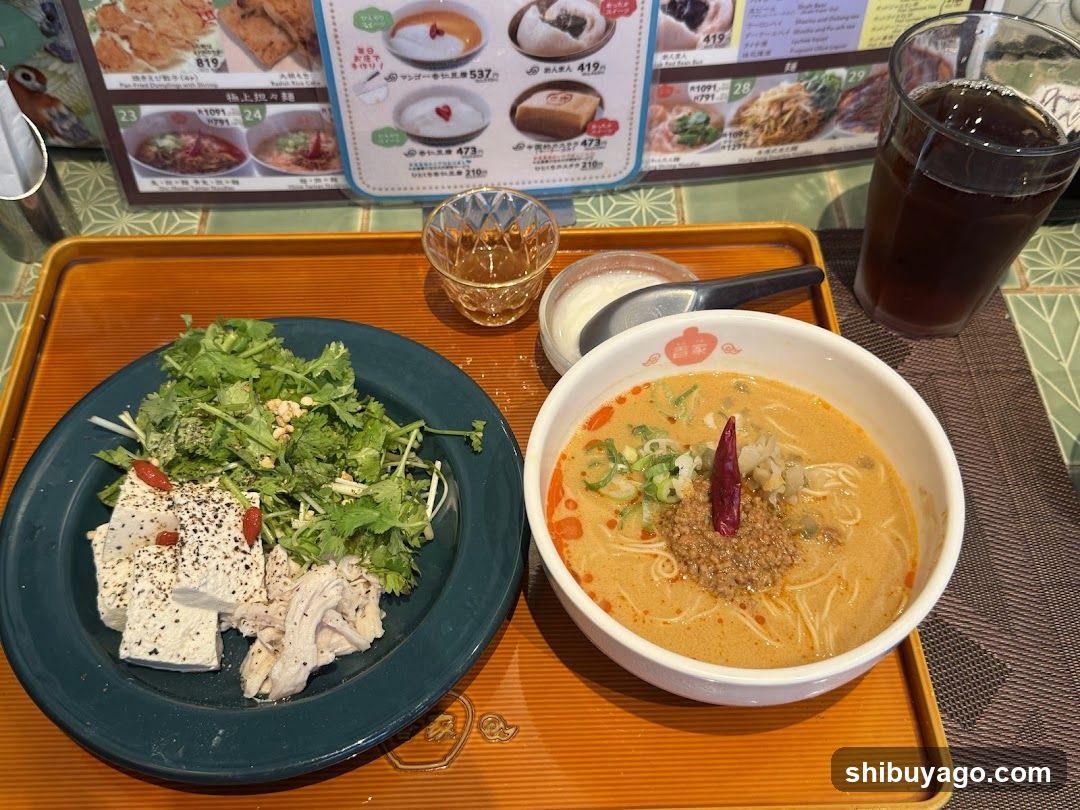 A tray with a bowl of spicy ramen topped with chili flakes, green onions, and a red chili pepper. Beside it is a plate with tofu cubes, shredded chicken, cilantro, and peanuts. A glass of dark soda and a small cup of sauce are also on the tray. In the background, a menu board displays various food items with prices.