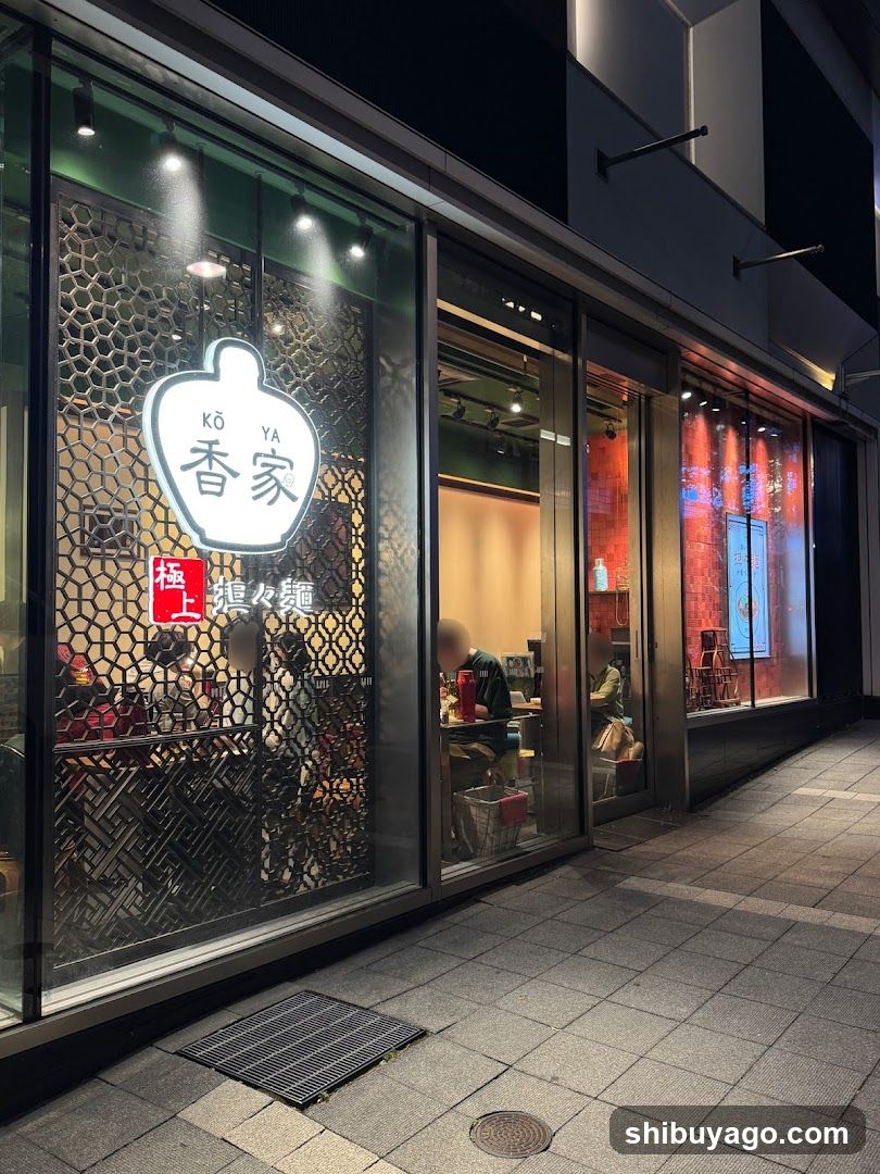 The image shows the exterior of a ramen shop named 'Koya' (香家) with illuminated signage on the glass facade. The entrance features large windows revealing the interior, where customers are seated at tables. A decorative lattice pattern is visible behind the counter area. The street outside appears quiet and well-lit by artificial lighting.