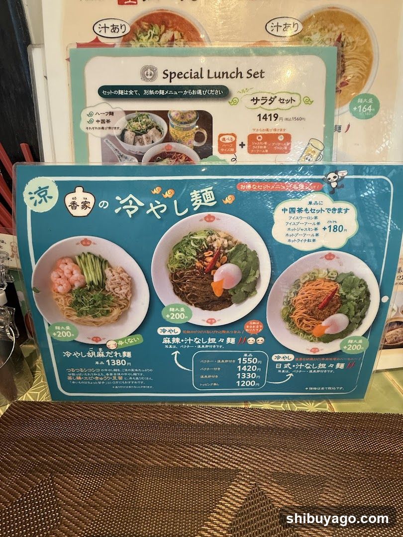 A close-up of a blue menu board featuring cold noodle dishes with prices and descriptions in Japanese, placed on a table next to another promotional card.