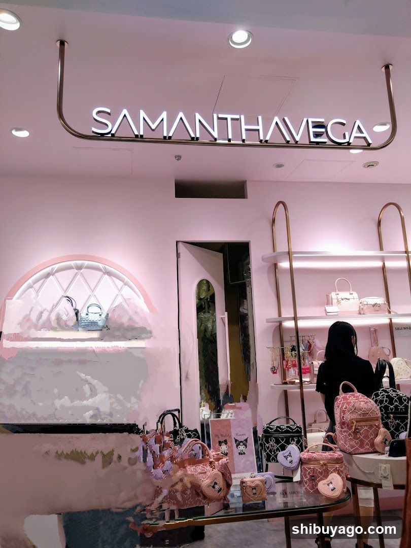 A pink-themed retail store with 'SAMANTHAVEGA' signage mounted on the ceiling above the entrance. The interior displays various handbags and accessories, including a glass table in the foreground holding several bags. A person is visible near the shelves on the right side of the image.
