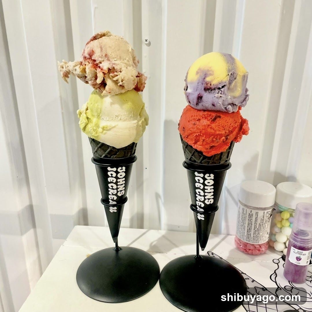 Two ice cream cones on black stands with 'JOHNS ICE CREAM' branding. Left cone has green and beige scoops topped with strawberry swirl. Right cone has orange, purple, and yellow scoops. Background shows white vertical blinds and jars of colorful candies.