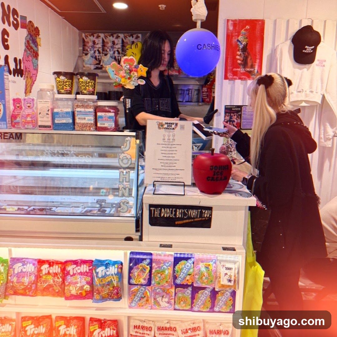 Interior of an ice cream shop with a counter, display cases, and merchandise. A staff member is serving a customer at the checkout area. Various branded candies are displayed on shelves below the counter.