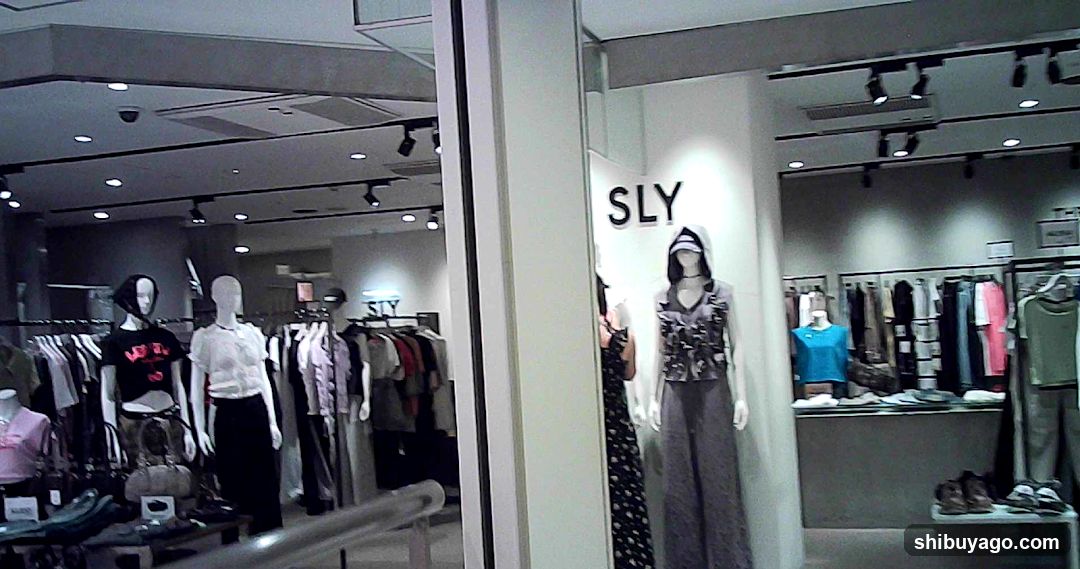 A view of a clothing store entrance, likely SLY based on the visible signage. The storefront features mannequins dressed in various outfits displayed both inside and outside the shop. Inside, racks of clothes are visible under track lighting.
