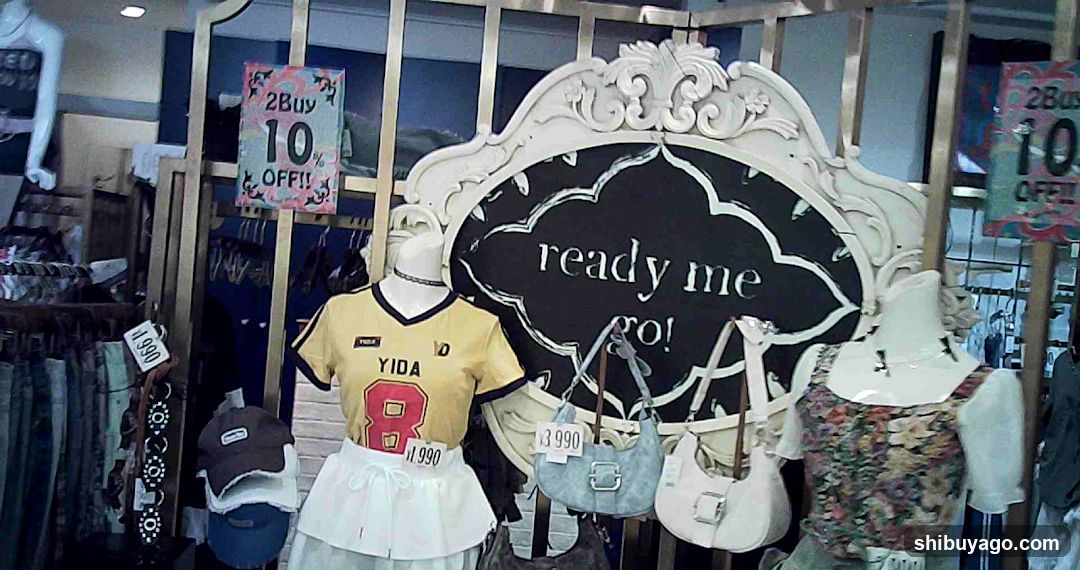 Storefront features a decorative white sign with 'ready me go!' text, mannequins wearing yellow jersey and floral dress outfits, hanging handbags priced at $3990, promotional signs offering '2 Buy 10 OFF', clothing racks visible inside, and partial view of another sale sign on the right.