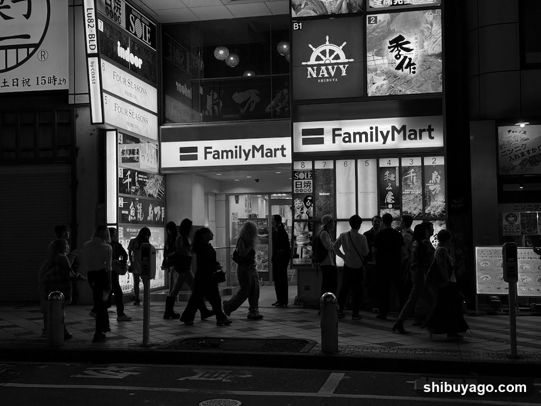 Black and white photo of a FamilyMart entrance with visible signage, illuminated by streetlights and shop lights. Pedestrians walk along the sidewalk in front.