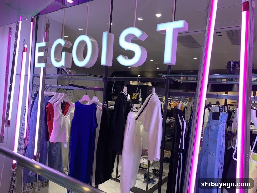 A modern clothing store entrance featuring a large illuminated sign reading 'EGOIST' in white block letters mounted above the glass facade. Vertical pink neon tubes frame the storefront on both sides, adding vibrant lighting. Inside, racks of women's fashion are visible, including denim jeans, dresses, and tops in various colors like blue, red, black, and white. The interior is well-lit with recessed ceiling lights.