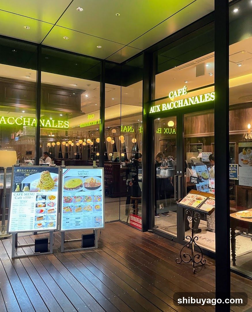 The image shows the exterior entrance of a café with glass doors and windows displaying illuminated signage reading 'CAFÉ AUX BACCHANALES'. Inside, warm lighting and wooden interiors are visible. Outside, two menu boards stand on the wooden deck near the entrance.