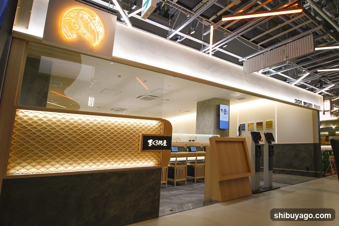 The image shows a modern shop entrance featuring an illuminated golden logo above the glass facade. Inside, there are wooden tables and chairs arranged neatly, along with digital screens displaying information. The interior is well-lit with warm lighting, creating a welcoming atmosphere. A signboard with Japanese characters is visible near the entrance, indicating the name of the establishment.
