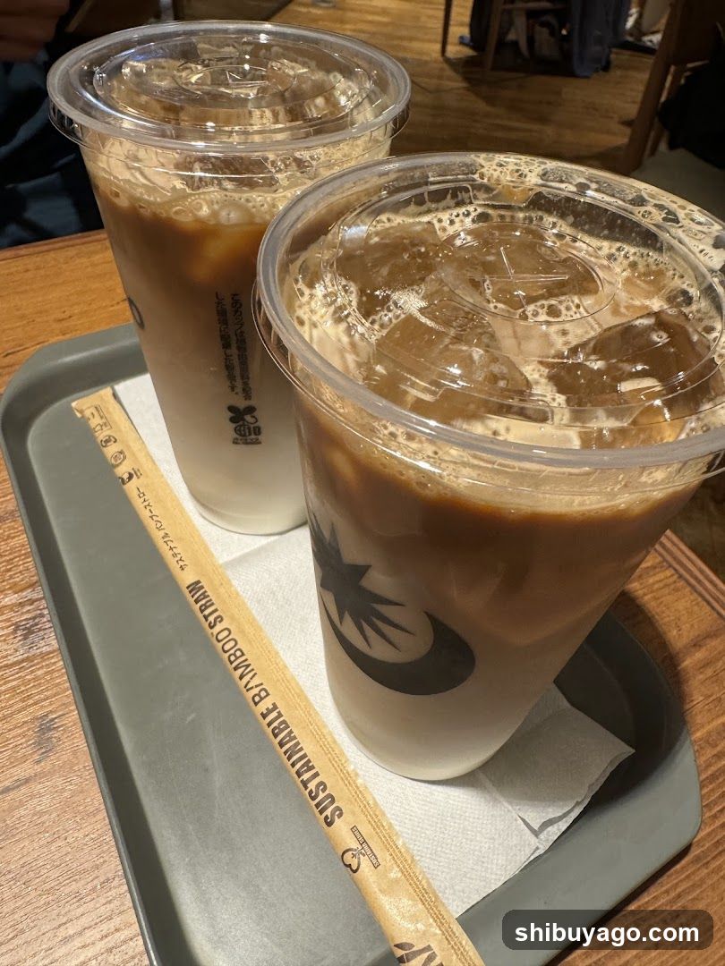 Two cups of iced coffee with ice cubes and foam, placed on a gray tray lined with paper napkins. A bamboo straw wrapper labeled 'SUSTAINABLE BAMBOO STRAW' is visible next to the cups. The cups have clear lids with logos.