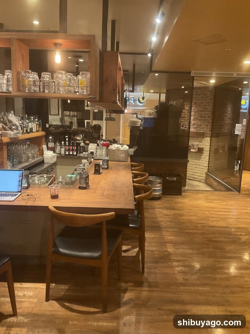The image shows the interior of a warmly lit establishment with wooden flooring and furniture. A long wooden counter runs along the left side, lined with several chairs. On the counter, there is a laptop, some glasses, and other small items. Above the counter, shelves hold various bottles and jars. In the background, a glass-enclosed kitchen area is visible, with brick walls adding to the rustic ambiance. The lighting is soft, creating a welcoming atmosphere.