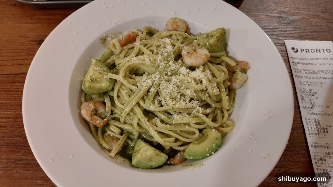 A plate of green pesto pasta topped with shrimp, avocado slices, and grated cheese, served on a wooden table next to a receipt from PRONTO.