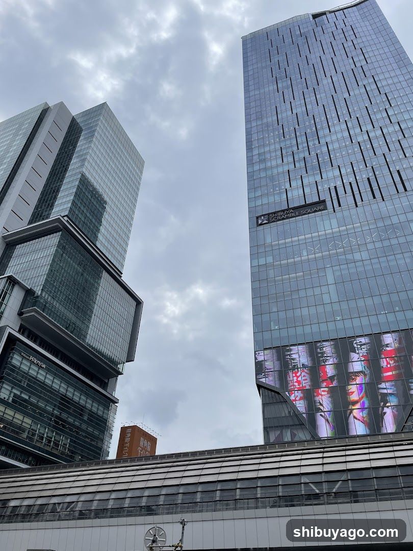 A low-angle view of modern glass skyscrapers under a cloudy sky. The building on the right features a large digital screen displaying colorful abstract visuals near its base. A lower structure with horizontal slats runs across the foreground, possibly part of a transit or commercial complex.
