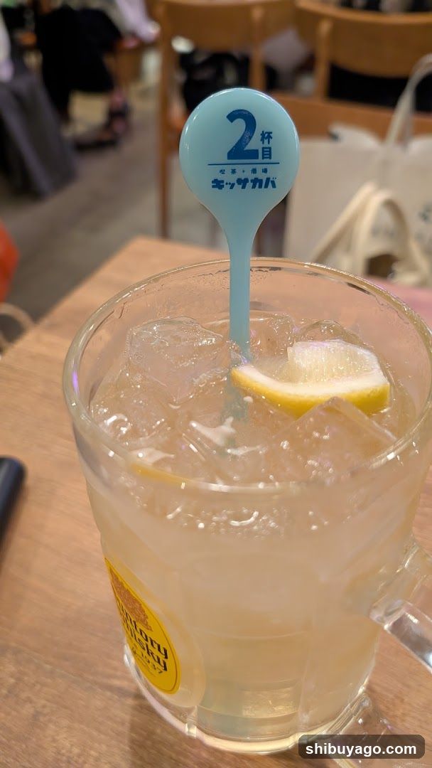 A glass mug filled with an icy lemonade drink, garnished with a lemon wedge and ice cubes. A blue stirrer stick labeled '2nd cup' is inserted into the drink. The setting appears to be indoors at a casual dining or cafe environment.