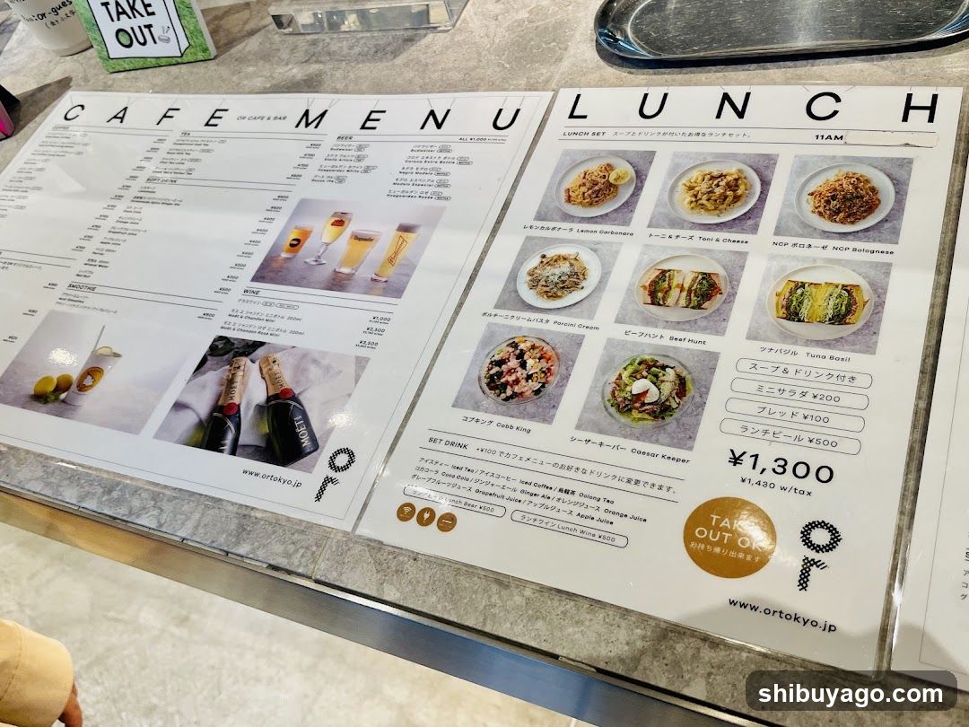 A close-up of a cafe menu spread on a table, featuring sections labeled 'CAFE & BAR' and 'LUNCH'. The lunch section lists items like Lemon Cornish Chicken, Tuna & Cheese, NCP Bolognese, Garlic Pasta, Beef Hunt, Tuna Bowl, Caesar Salad, and Cock Ring. Prices are listed in yen, with a set drink option at ¥1,300 including tax. A 'TAKE OUT' sign is visible in the background.