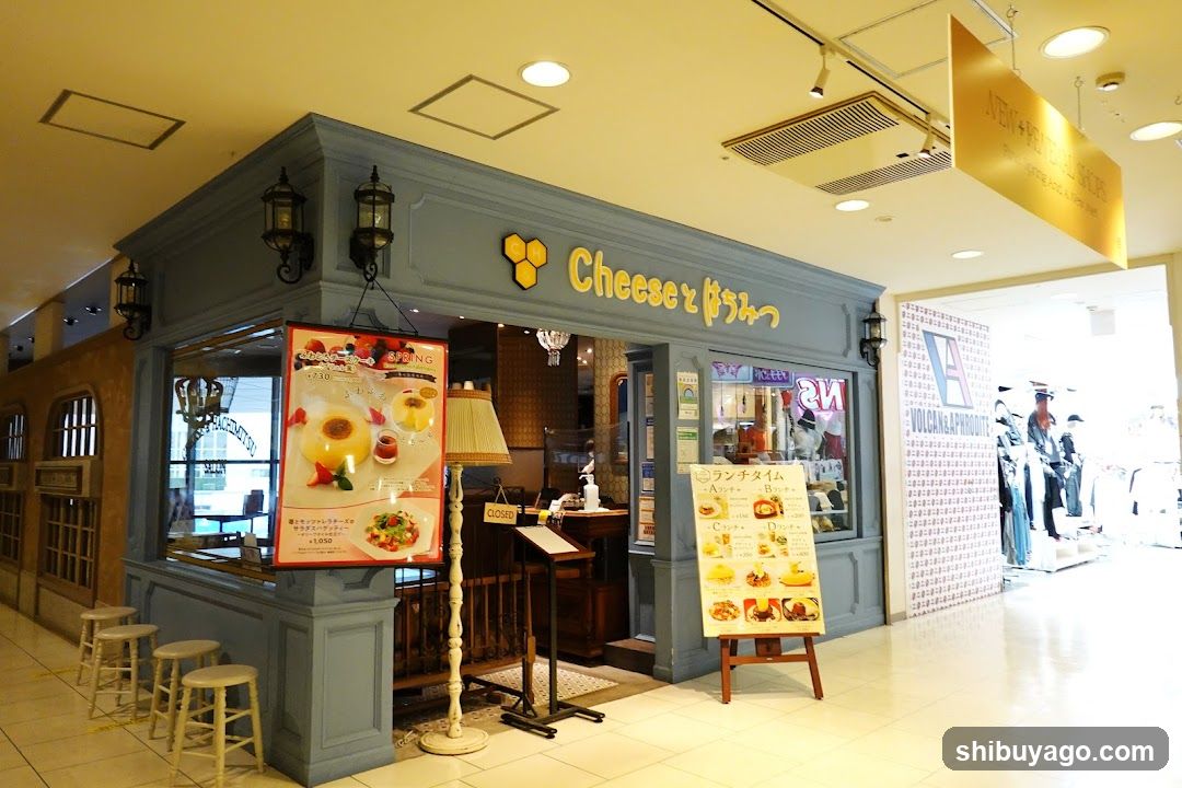 A blue-painted shop front with a yellow honeycomb logo and Japanese text above the entrance. A large poster on the left window displays food items, including desserts and sandwiches. Inside, there is a counter, stools, and a lamp. Outside, a menu board stands near the entrance showing various food options with prices.