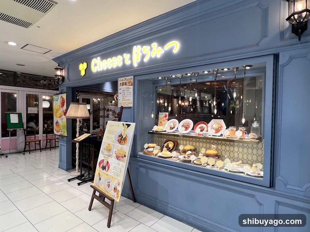 A blue-painted shop facade with illuminated neon signage reading 'Cheese と ばらみつ'. The window display showcases various pastries and desserts on shelves, including cakes, tarts, and small cakes. A menu board stands outside the entrance, featuring images of food items. Inside, hanging lights illuminate the interior space.
