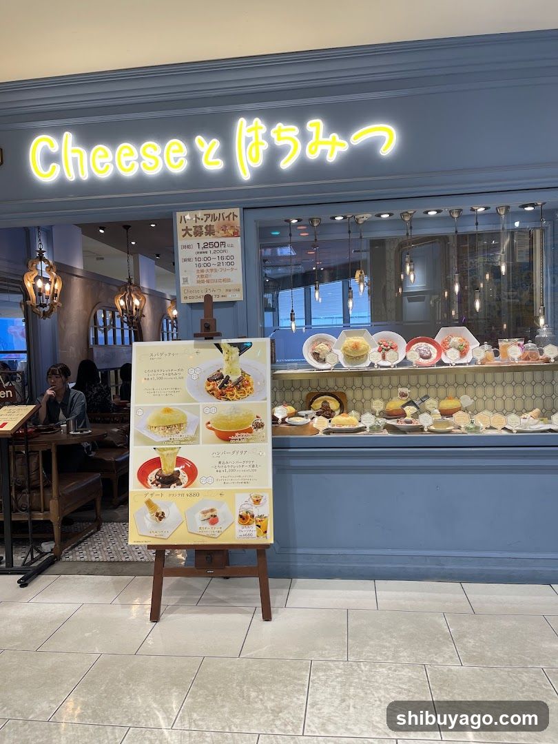 A blue storefront with illuminated yellow neon sign reading 'Cheese とぼちみつ'. Inside, a display case showcases various food items including desserts and pastries. A menu board stands outside the entrance.