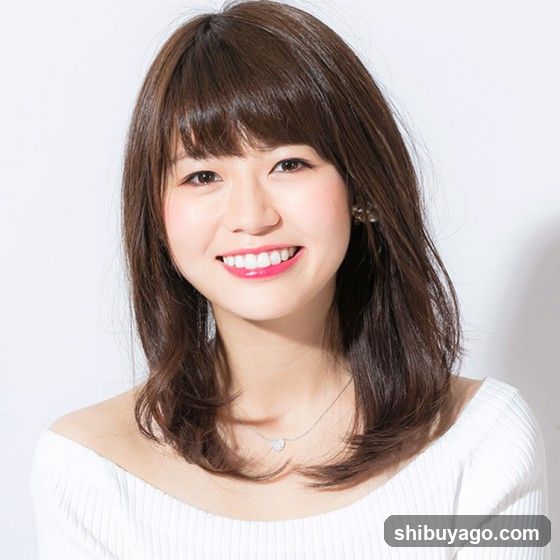 Portrait of a smiling woman with shoulder-length brown hair and bangs, wearing a white off-the-shoulder top and a delicate necklace.
