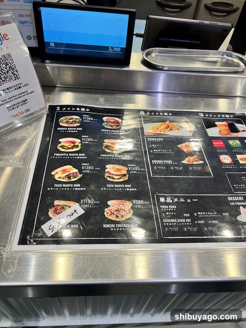 A close-up of a food menu board displayed on a stainless steel counter, featuring various sandwich options and side dishes with prices in yen.