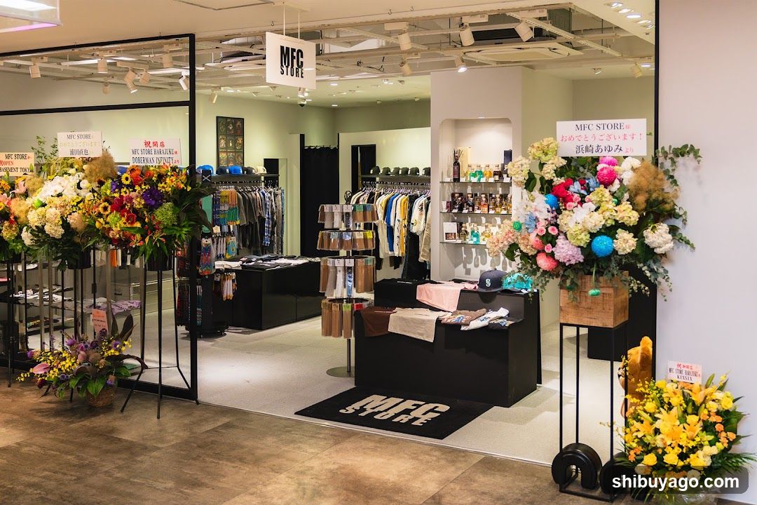 The image shows the entrance of an MFC STORE, featuring a clean and modern interior design. The store is adorned with vibrant floral arrangements on both sides of the entrance, adding a welcoming touch. Inside, racks of clothing are neatly arranged, showcasing a variety of garments. A sign above the entrance reads 'MFC STORE,' confirming the store's identity. The lighting is bright, highlighting the products and creating an inviting atmosphere.