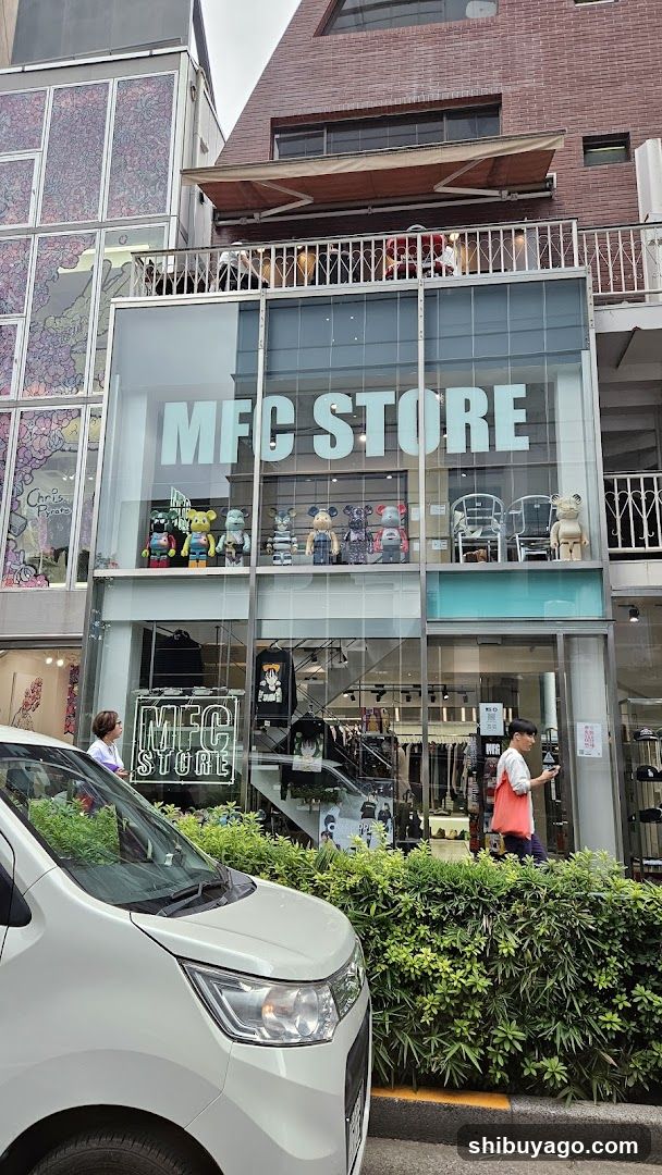 A modern glass-fronted storefront with large 'MFC STORE' signage on the upper facade and a smaller illuminated sign at ground level. The shop displays various merchandise including plush toys, clothing racks inside, and decorative items on an upper balcony. A white van is parked in front, partially obscuring the lower view. Green shrubbery lines the sidewalk.