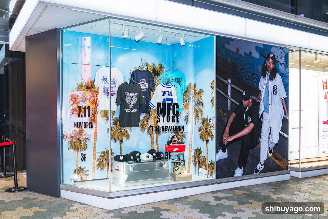 A brightly lit storefront window featuring a vibrant blue backdrop with palm trees and the '711' logo. Inside, various clothing items including t-shirts and jerseys are displayed on hangers. A red shoe box sits atop a metal trunk in the foreground. To the right, two individuals pose against a mural-style wall. The scene suggests a new retail opening at level 3F.