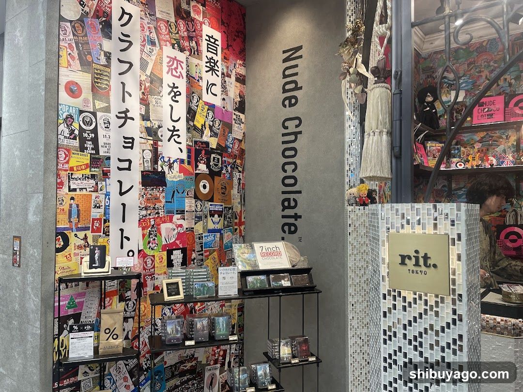 Store entrance with 'Nude Chocolate' vertical signage on gray wall, mosaic-tiled pillar displaying 'rit. TOKYO', and colorful pop-art collage background. Shelves hold packaged goods and framed items.