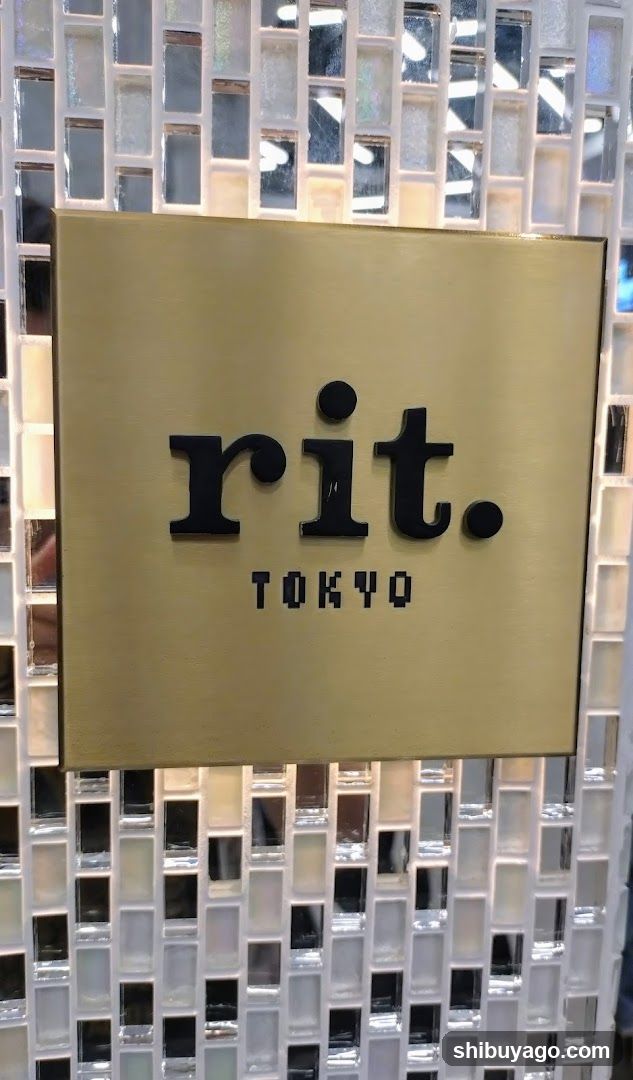 A close-up photo of a gold-colored square sign mounted on a glass wall with mosaic tiles. The sign displays the word 'rit.' in lowercase black letters, followed by a period and the word 'TOKYO' below it in pixelated font.